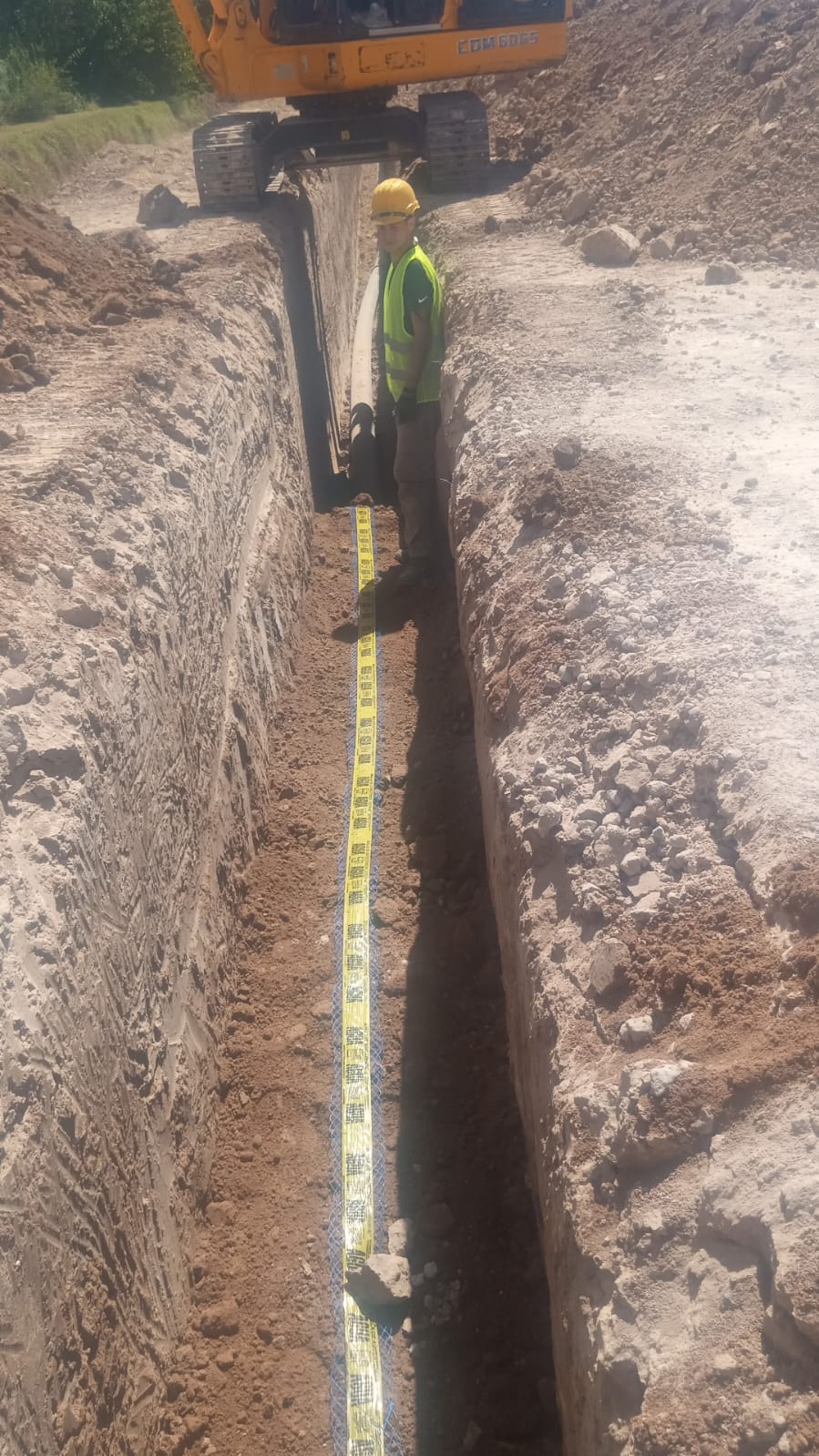 Trabajador con equipo de protección personal junto a una zanja abierta para tendido de cañería PEAD, con retroexcavadora en el fondo.